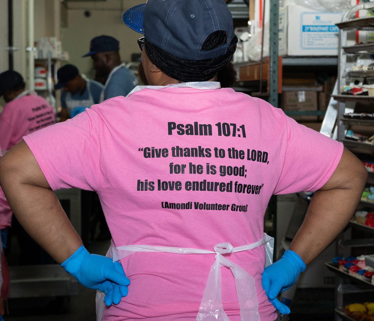 A volunteer seen from behind in a pink T-shirt printed with Psalm 107:1 and Amondi Volunteer Group, in a kitchen with other volunteers